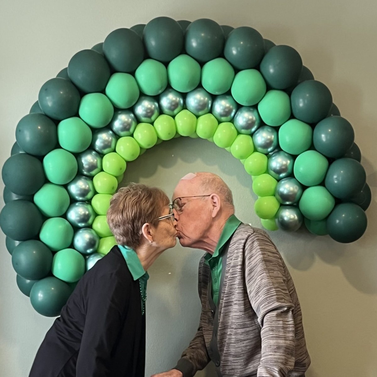 A married couple sharing a kiss beneath a green balloon arch at a community celebration